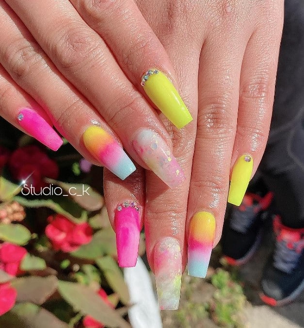 A closeup of a woman's long coffin nails with yellow and pink nail polish that has glitter design and a gradient effect in yellow, pink, and blue and tiny studs near the cuticles