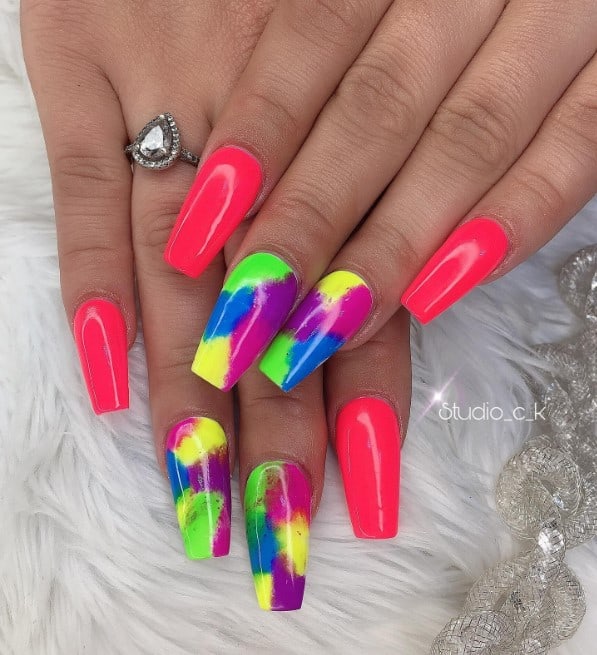 A closeup of a woman's long nails with bright fuschia pink manicure that has smudges of vivid colors like green, blue, violet, magenta, and yellow on accent nails 