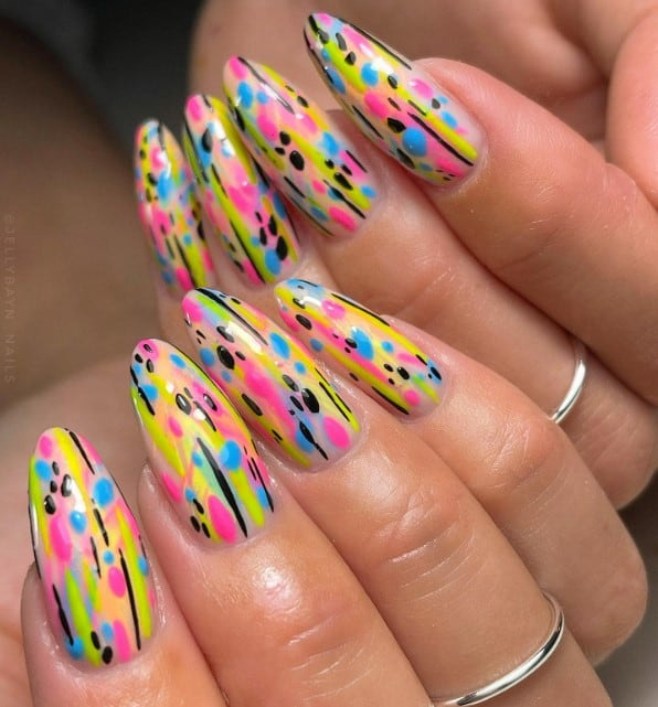 A closeup of a woman's long almond nails with pink and blue dots, short black lines, and streaks in assorted bright colors