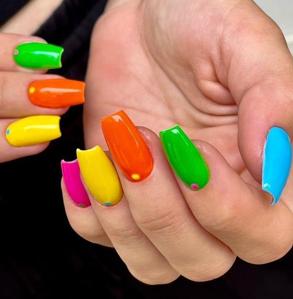 A closeup of a woman's long coffin nails with vibrant colors like green, orange, yellow, pink, and blue nail polish that has a tiny dot near the cuticles