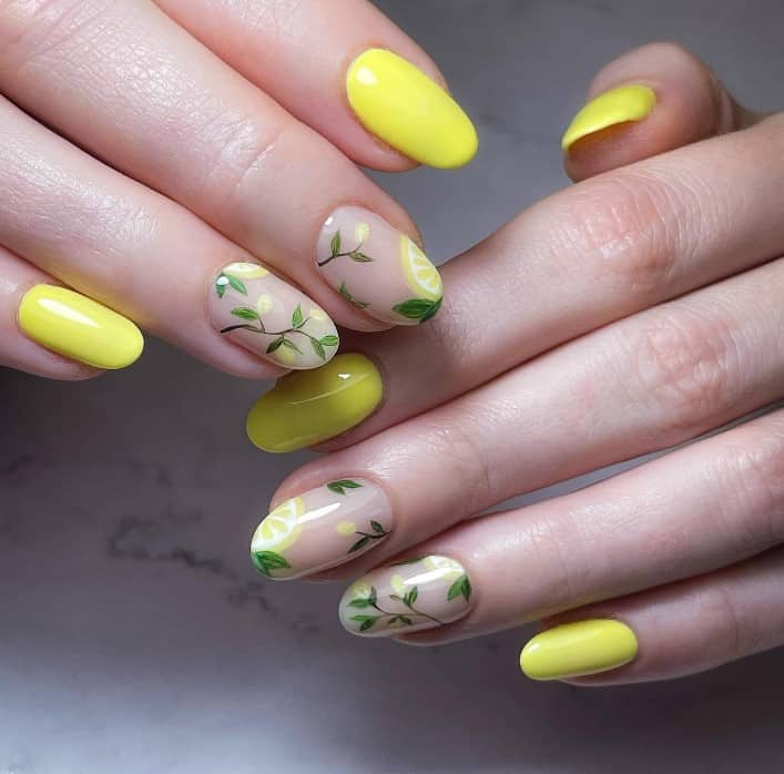 A closeup of a woman's fingernails with a nude and yellow nail polish base that has lemon branches and sliced lemons on select nails 
