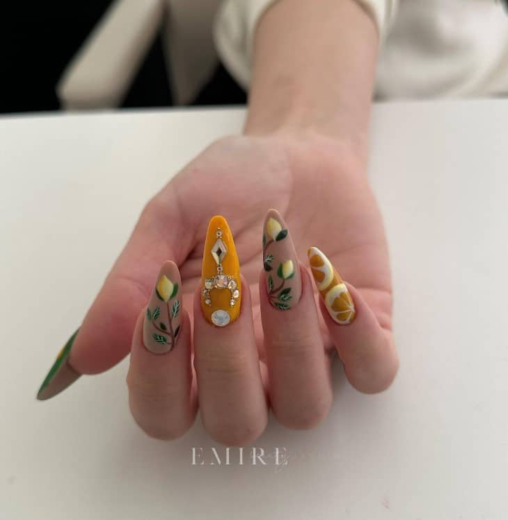 A woman's fingernails with yellow, beige, and green nail polish that has lemon slices, lemon fruits on branches, and dazzling jewels nail designs