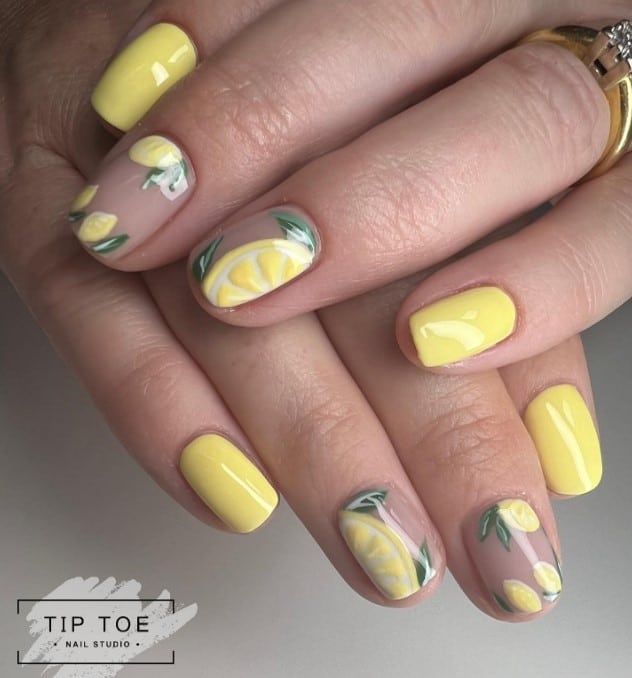 A closeup of a woman's fingernails with a nude and yellow nail polish base that has tiny lemons and giant sliced lemons occupying the nude accent nails