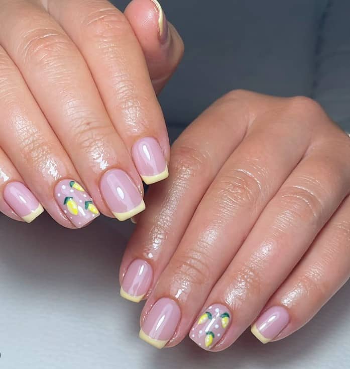 A closeup of a woman's fingernails with a glossy nude nail polish that has pale yellow French tips and an accent nail with lemon and polka dot decoration