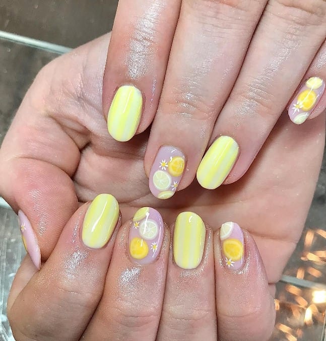 A closeup of a woman's fingernails with a combination of two shades of yellow in diagonal wallpaper-like strips on two nails that has lemon slices and lemon flowers on a milky white base