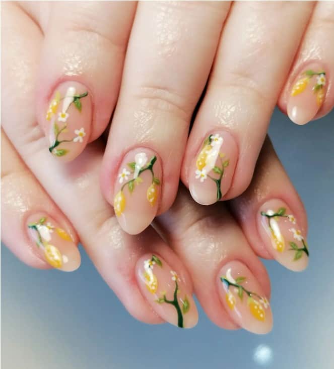 A closeup of a woman's long oval fingernails with   a sheer peach nail polish that has lemon tree branches with flowers
