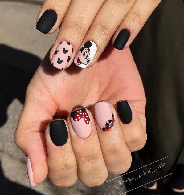 A woman's fingernails with a combination of matte black, baby pink, and white nails that has Mickey Mouse and Minnie Mouse art, iconic Mickey Mouse silhouettes, and rhinestone cuticle cuffs in black, red, and silver
