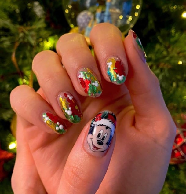 A closeup of a woman's fingernails with clear base that has red, green, and gold foil flakes and white Mickey silhouettes and Mickey Mouse in a Santa hat on select nails