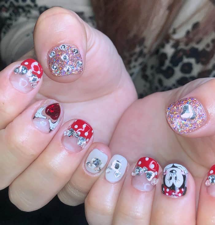 A woman's fingernails with a clear nail base that has Christmas-themed Minnie Mouse nails with heart-, ribbon-, and Mickey-shaped rhinestones in silver and red, Mickey hands and more