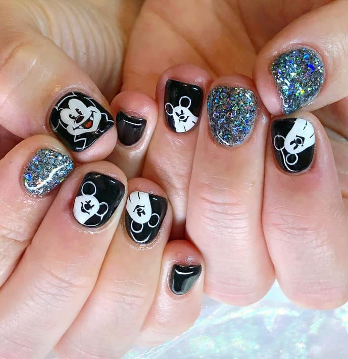 A closeup of a woman's fingernails with glossy black nails and silver glitter nails that has Mickey Mouse designs, Mickey Mouse designs and ghost Mickey art