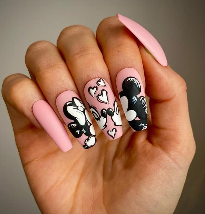 A woman's fingernails with matte pink nail polish base that has Mickey and Minnie Mouse nail art in black and white on select nails