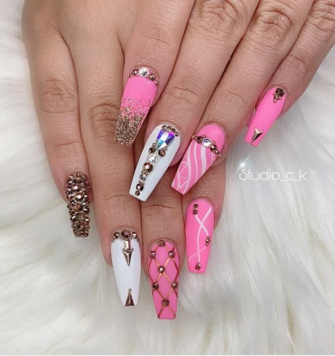A woman's coffin fingernails with a combination of white and pink nail polish base that has rhinestones, glitter, and jewels in different arrangements, crisscrossed patterns, swirls, and glitter ombré