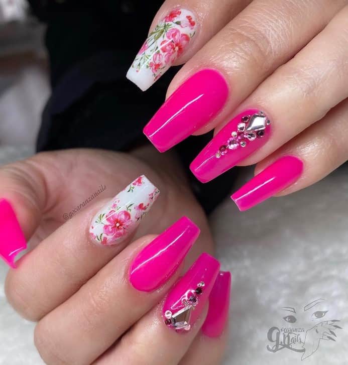 A closeup of a woman's coffin-shaped fingernails with neon pink and white nail polish that has flowers and gems on select nails 