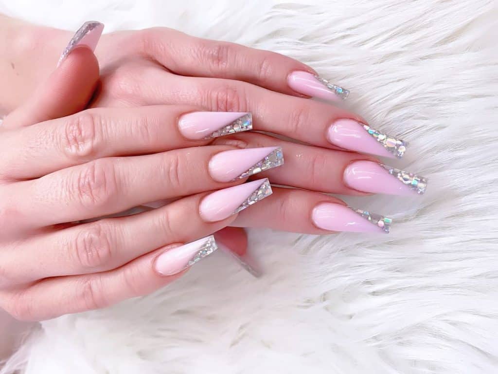 A closeup of a woman's fingernails with a light pink nail polish base that has diagonal rhinestones on nail tips 