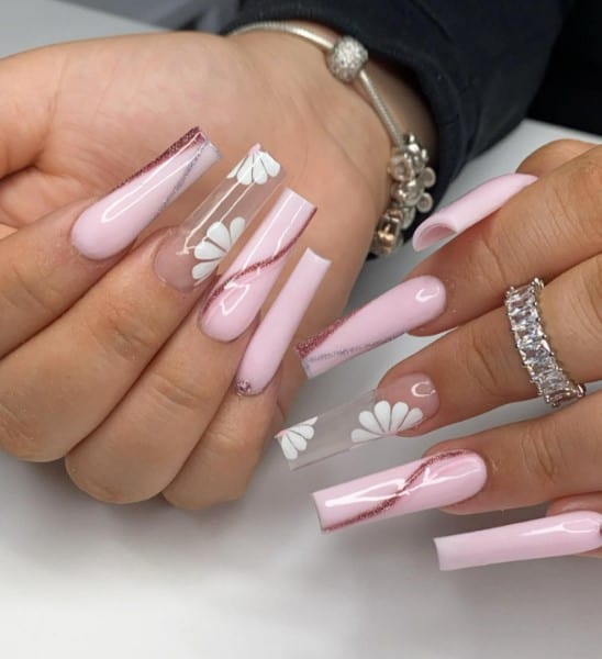 A closeup of a woman's coffin fingernails with pink acrylic nails that has swirls and two-toned French tips created with rose gold and silver glitter and clear accent nails with white flowers