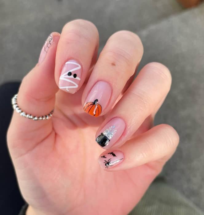 A closeup of a woman's fingernails with a nude nail polish base that has spiders, webs, a mummy, a cauldron and an orange pumpkin tip in the middle