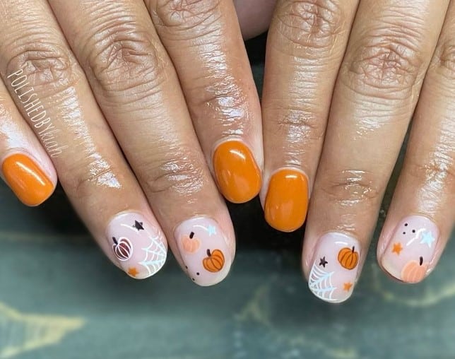 A closeup of a woman's fingernails with a glossy bright orange and white base that has a star, spiderweb, and pumpkin nail art