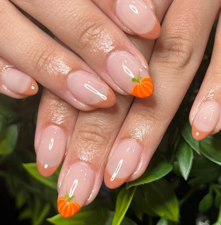 A closeup of a woman's fingernails with a glossy nude nail polish base that has light brown French tips and an orange pumpkin-shaped tip to one accent nail