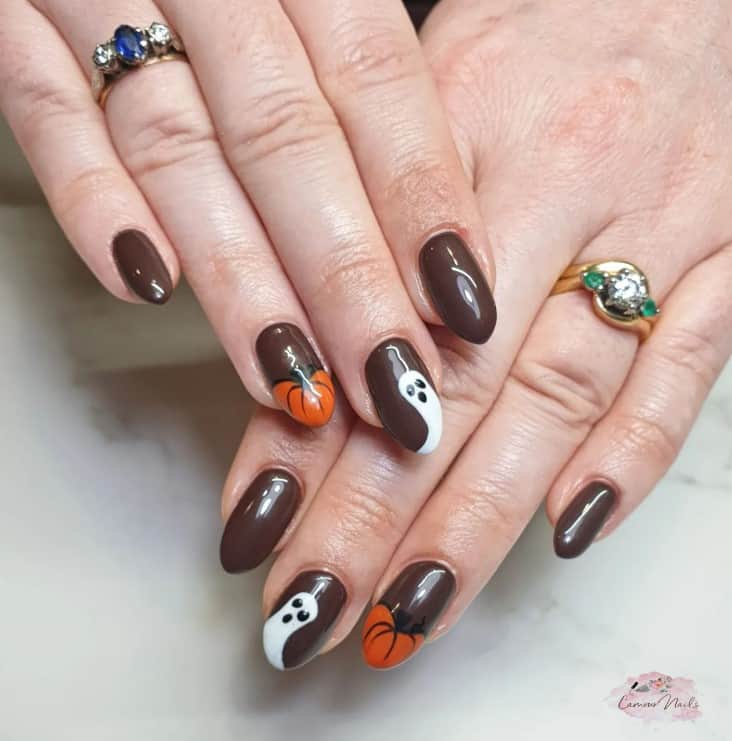 A closeup of a woman's fingernails with a brown nail polish base that has ghost and pumpkin on the tips