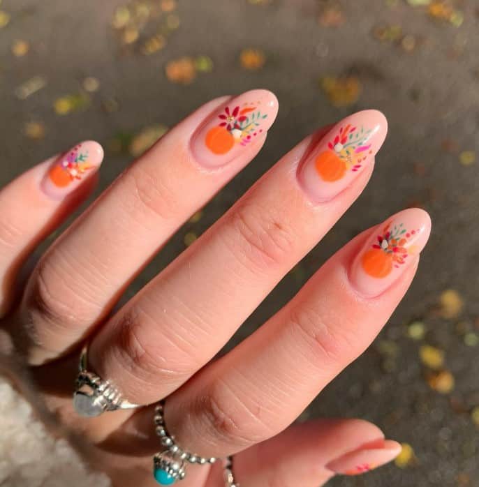 A woman's fingernails with a nude pink nail polish base that has colorful flowers, leaves, and pumpkins nail designs