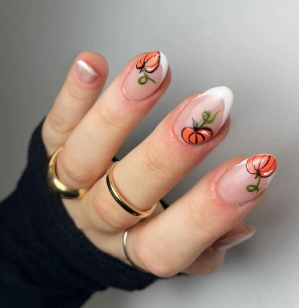 A woman's fingernails with a nude nail polish base that has pumpkins nail designs on select nails and white French tips 