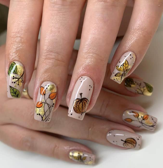 A closeup of a woman's fingernails with a light nude nail polish base that has hand-painted pumpkins, gold foil and fall leaves