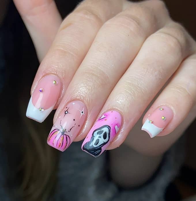 A closeup of a woman's fingernails with a pink and clear nail polish base that has Ghostface nail art, pink pumpkins, and solid white tips with sparkling gems