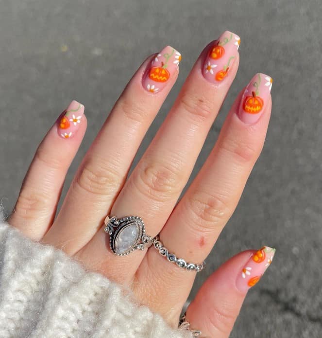 A woman's fingernails with a nude nail polish base that has jack-o’-lanterns and flowers nail designs
