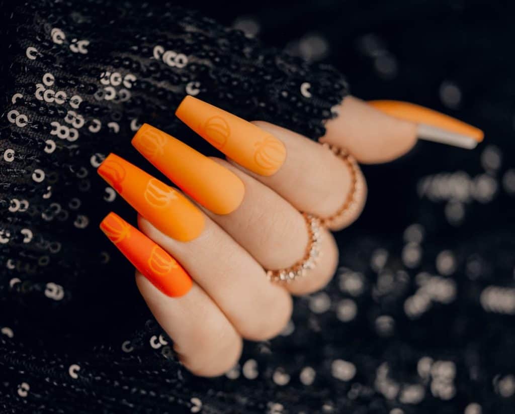 A woman's fingernails with matte orange nail polish in an ombre effect that has  embossed orange pumpkin designs in a shiny finish
