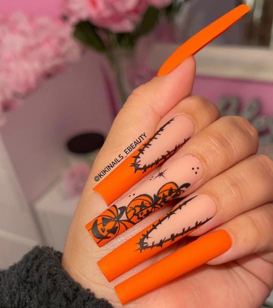 A closeup of a woman's fingernails with a mix of nude and matte orange nail polish that has jack-o’-lanterns and thorny outlines and matte orange French tips