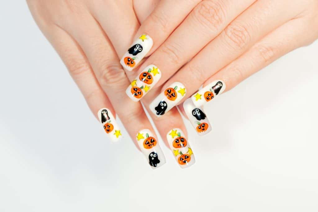 A woman's fingernails with a white nail polish base that has orange pumpkins and ghosts nail designs
