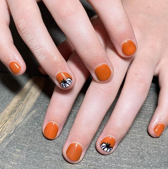 A closeup of a woman's fingernails with a solid orange nail polish base that has white pumpkin nail art on select nails 