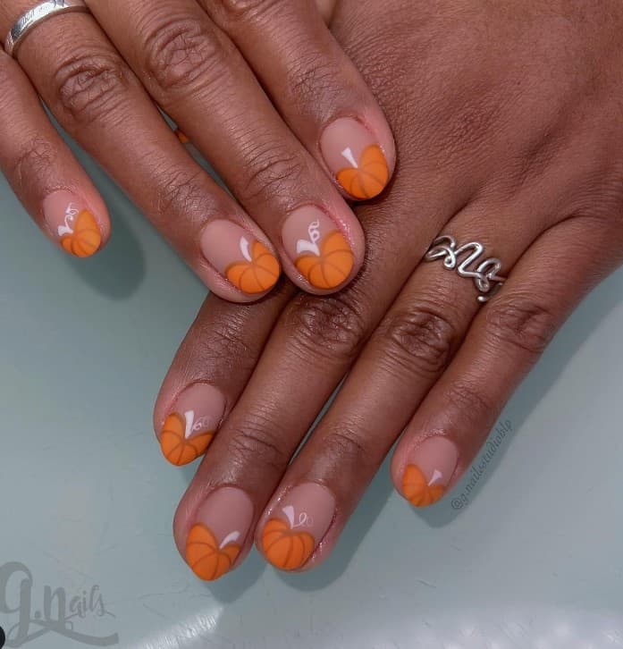 A closeup of a woman's fingernails with a nude nail polish base that has cute orange pumpkins on nail tips 