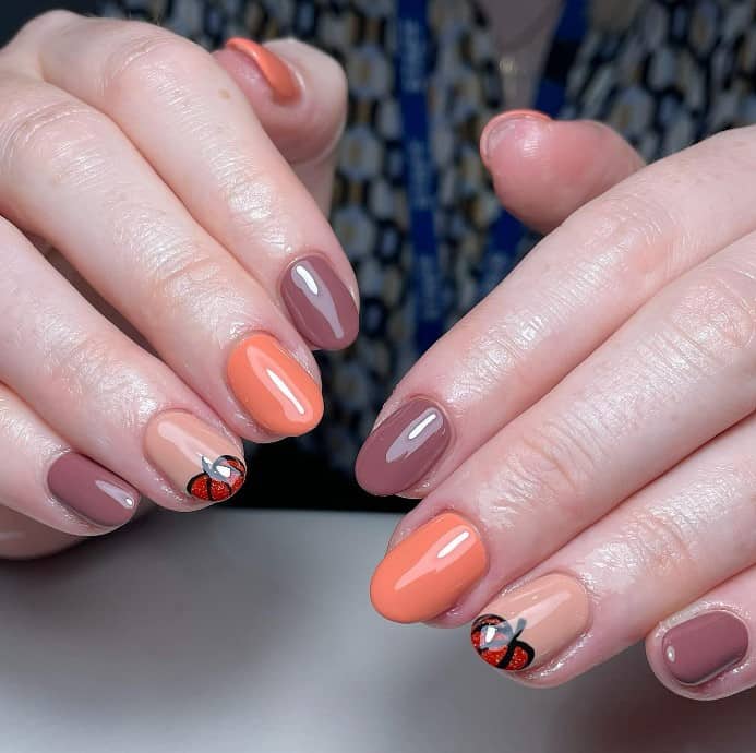 A closeup of a woman's fingernails with a mix of nude, peach, and brown nail polish base that has a mini pumpkin on the accent nails