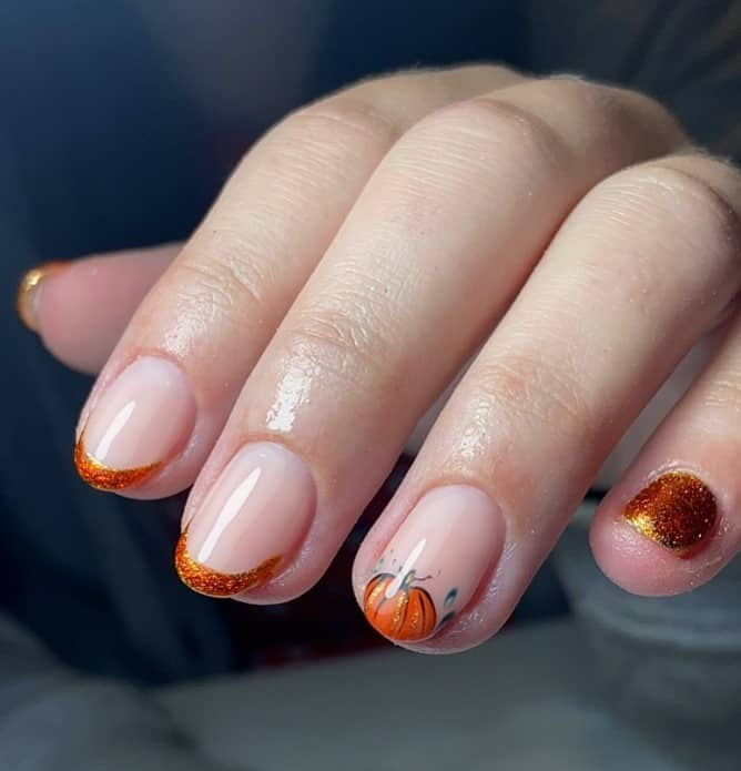 A closeup of a woman's fingernails with a nude nail polish base that has orange French tips, pumpkin nail tips and an orange glitter accent nails 