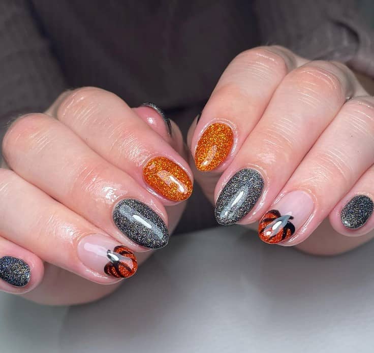 A closeup of a woman's fingernails with orange and black nails in glittery finish that has a small pumpkin on one nail