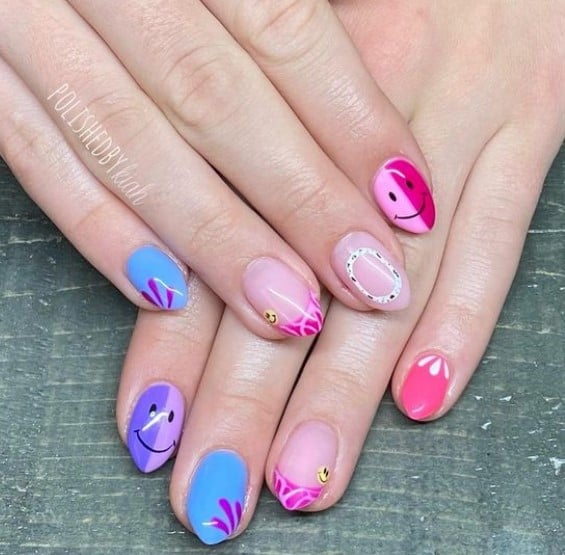 A closeup of a woman's nails with blue, pink, and purple colors that has patterned French tips, smileys, abstract lines, and color-block designs