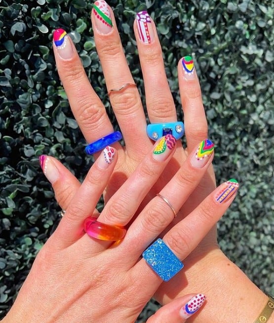 A woman's nude nails with different colors in varied patterns like animal prints, multicolored lines and dots, summer fruits, and multicolored V shapes on each nail