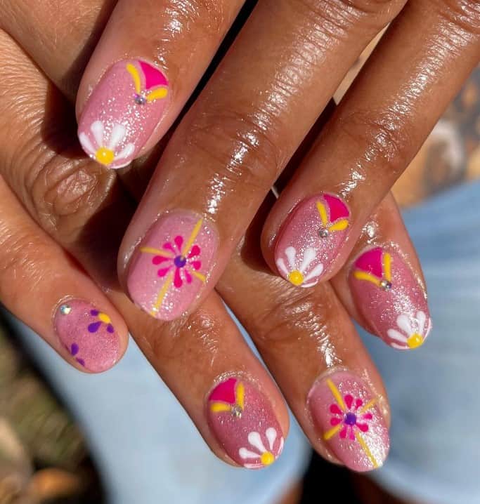 A closeup of a woman's nails with shimmery pink base that has deep pink, purple, white, and yellow floral designs