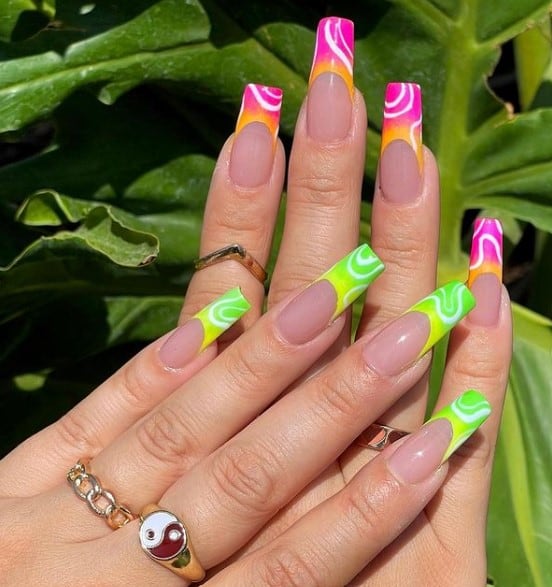 A closeup of a woman's long nails with nude nail polish base that has ombré French tips in hot pink and orange on the nails of one hand and lime green and yellow on the other with white swirls