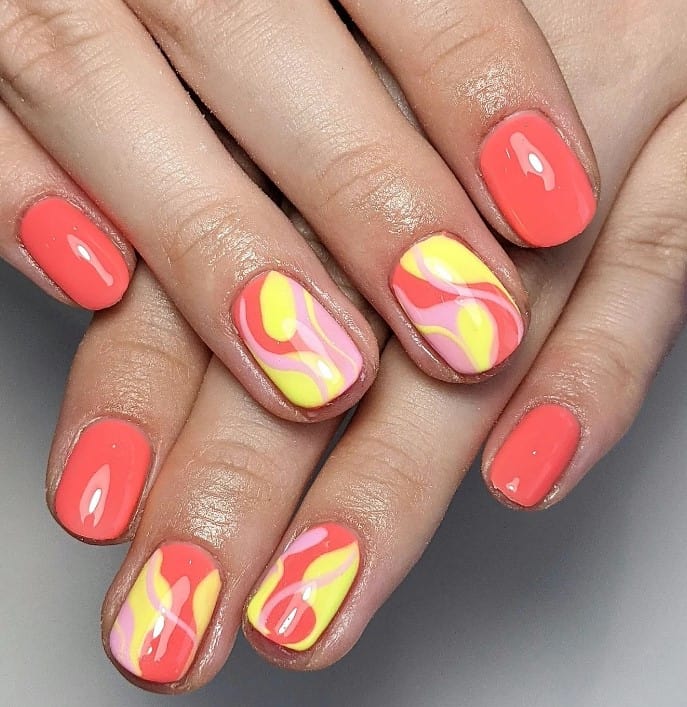 A closeup of a woman's short nails with coral and sunny yellow nail polish that has swirl accents