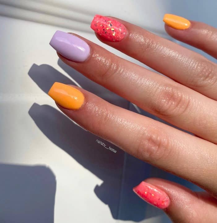 A closeup of a woman's short nails with orange, purple and salmon pink nail polish base that has metallic glitter on pink nails