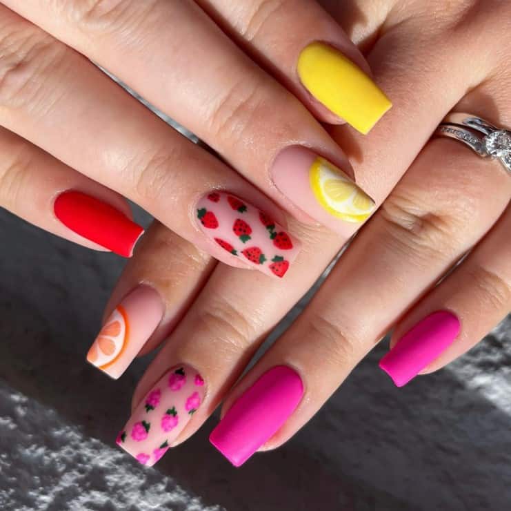 A closeup of a woman's nails with fuschia pink, bright yellow, and red, combined with accent nails that has strawberries, raspberries, and lemon and orange slices