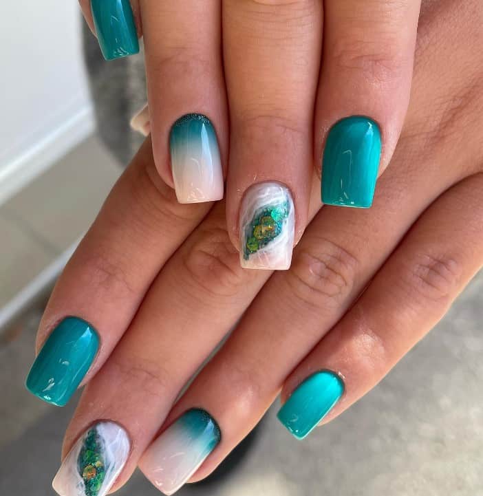 A closeup of a woman's mid-length nail with deep teal to crisp white nail polish in an ombre effect that has an intricate marble design with glittery waves