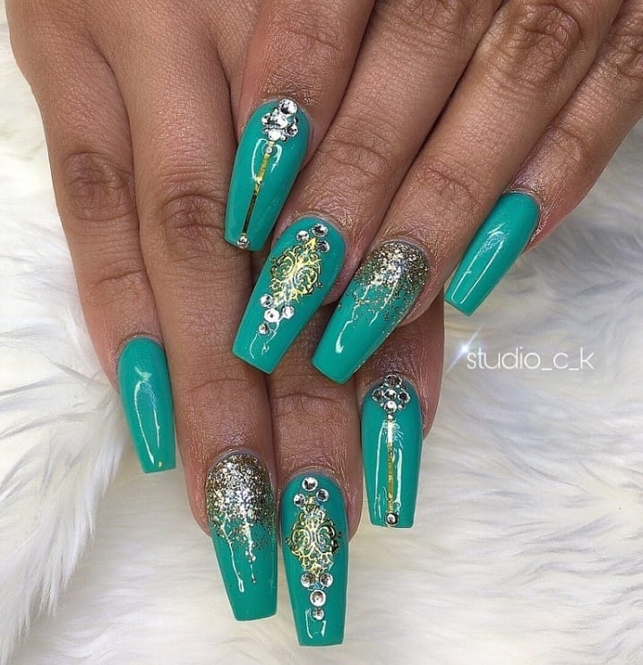 A closeup of a woman's long nails with teal nail polish that has ornate gold accents, glitter ombre, gold lines, and rhinestones nail designs