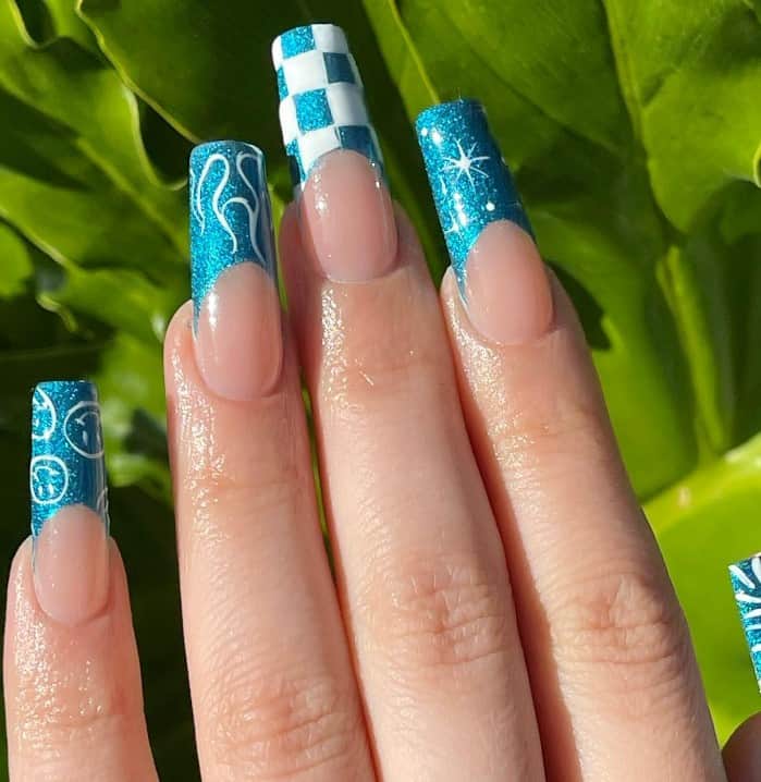 A closeup of a woman's fingernails with long square nails that have glittery teal French tips that has smileys, swirls, stars, and playful checkered patterns