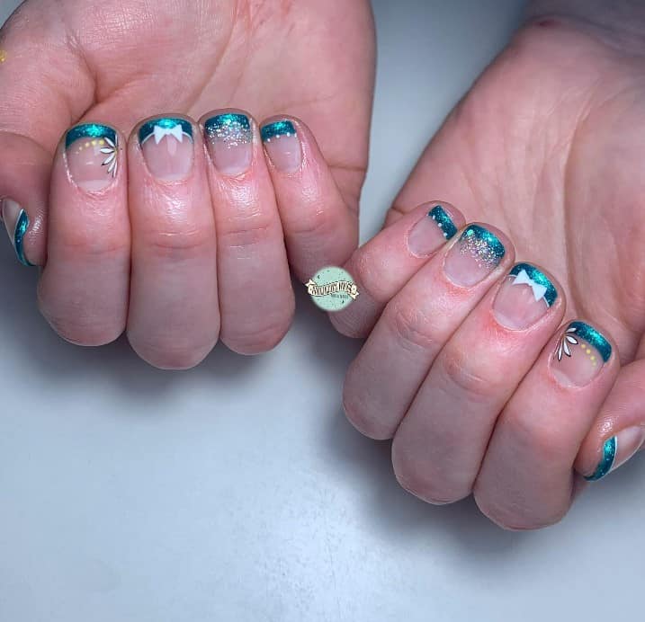 A closeup of a woman's short nails with nude nail polish base that has glittery teal French tips decorated with white dots, glitter flakes, intricate flower petals, and charming ribbon accents
