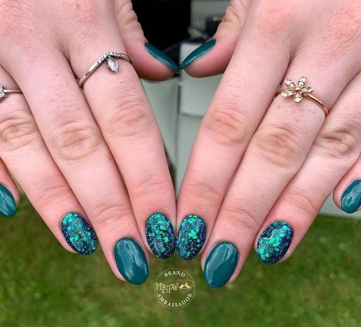 A closeup of a woman's short nails with teal nail polish base that has small glitter particles and oversized glitter flakes  on select nails 