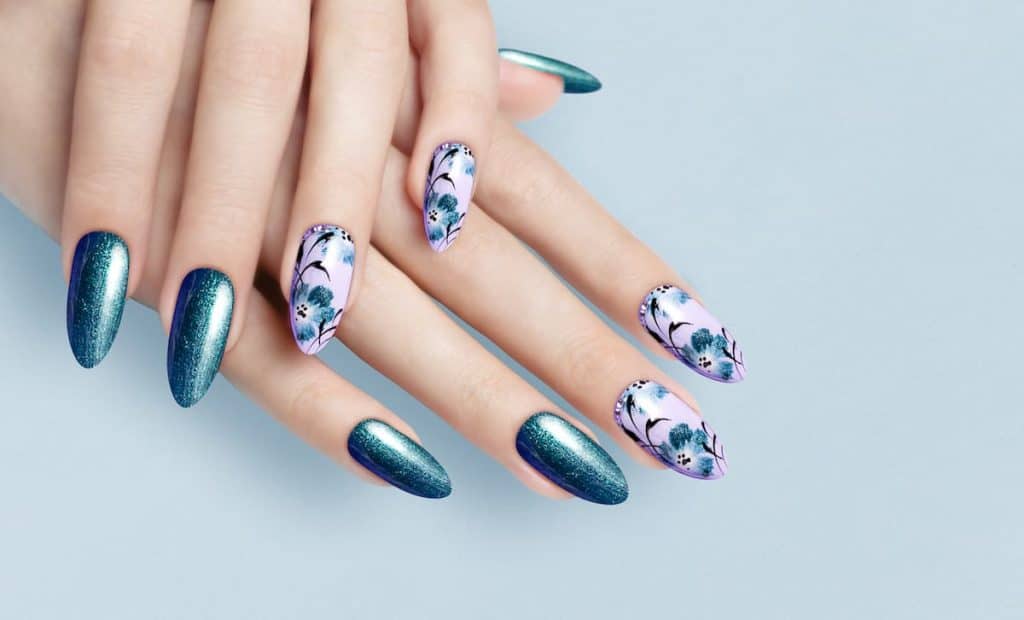 A woman's long nails with purple and glittery teal nail polish that has floral nail designs on purple nails 
