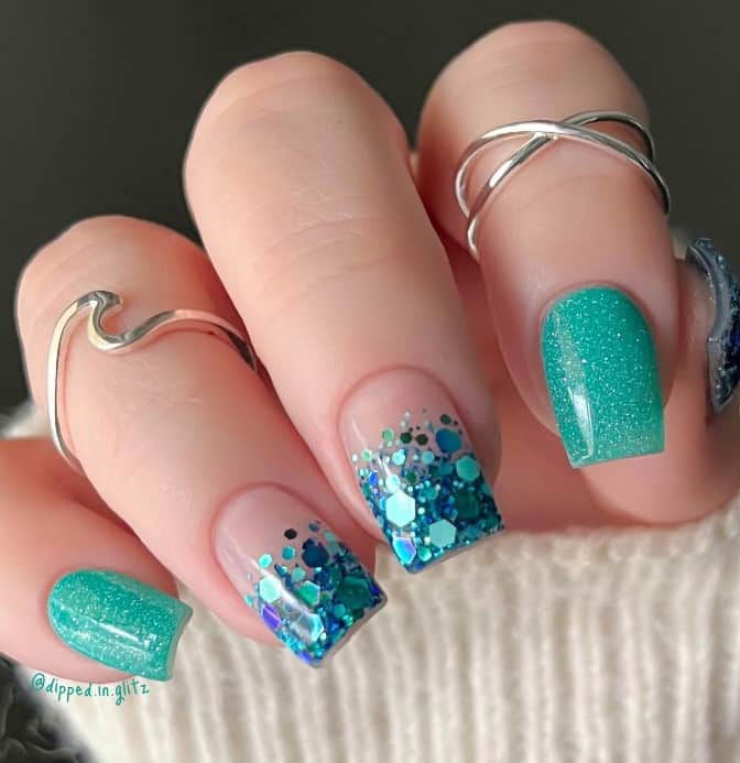 A closeup of a woman's long nails with nude and teal glitter nail polish that has addition of confetti-like blue and teal glitter flakes in an ombre effect on nude nails 